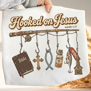 May include: White graphic tee with the text "Hooked on Jesus" and Mark 1:17. The design features a fishing rod with various religious symbols hanging from hooks, including a Bible, cross, and fish.