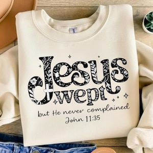 May include: Cream-colored sweatshirt with the words "Jesus wept" in a black floral design. The text "but He never complained John 11:35" is below the main text. The sweatshirt is folded, with a hat and jeans visible in the background.