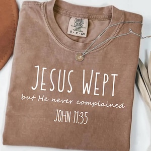 May include: A brown Comfort Colors t-shirt with the text "JESUS WEPT but He never complained JOHN 11:35" in white. A silver necklace with a small pendant rests on the shirt.