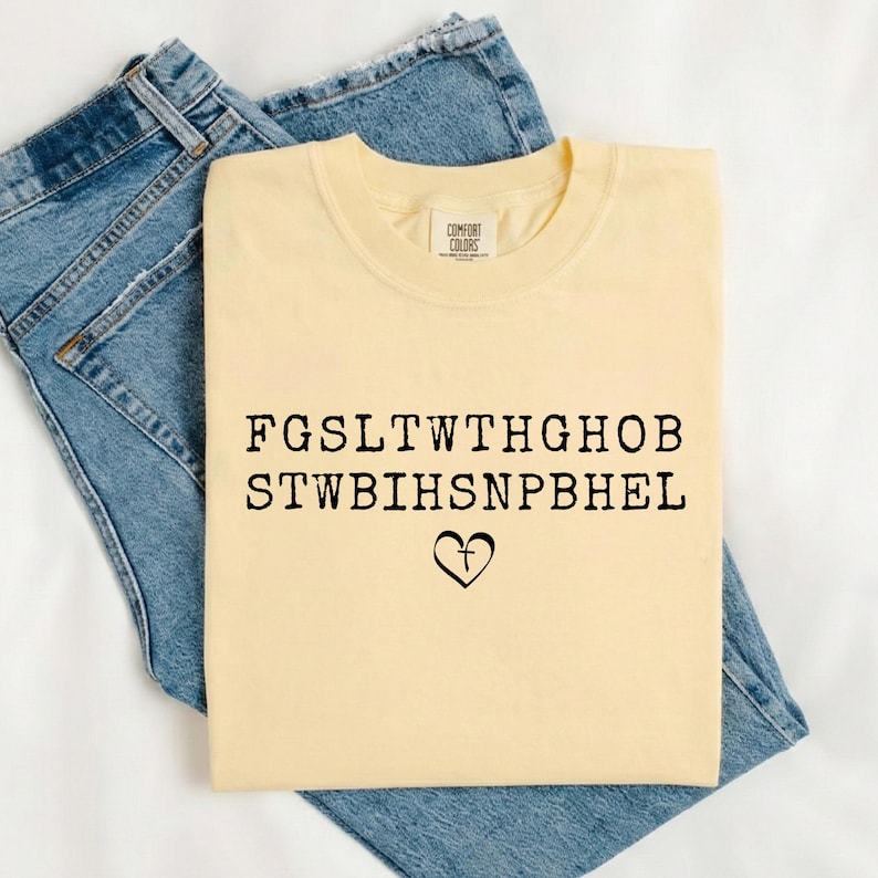 May include: A pale yellow Comfort Colors t-shirt featuring black text and a heart with a cross graphic, folded on blue denim jeans. The text is in a bold, sans-serif font. The t-shirt has a Comfort Colors label.