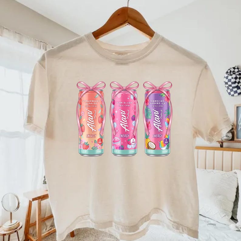 May include: A cream-colored t-shirt displays three Alani Nu energy drink cans. The cans are decorated with pink bows and feature colorful designs: Strawberry Sunrise, Pink Slush, and Hawaiian Shaved Ice. The Alani Nu logo is visible on each can.