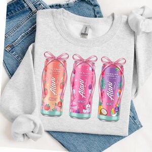 May include: A light gray sweatshirt featuring three colorful Alani Nu energy drink cans. The cans are decorated with pink bows and flip-flop straps. Each can has a different flavor: Strawberry Sunrise, Pink Slush, and Hawaiian Shaved Ice.