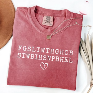 May include: A dusty rose Comfort Colors t-shirt with white text and a heart with a cross symbol. A gold necklace with a pendant rests on the shirt. The shirt is folded and has a tag.