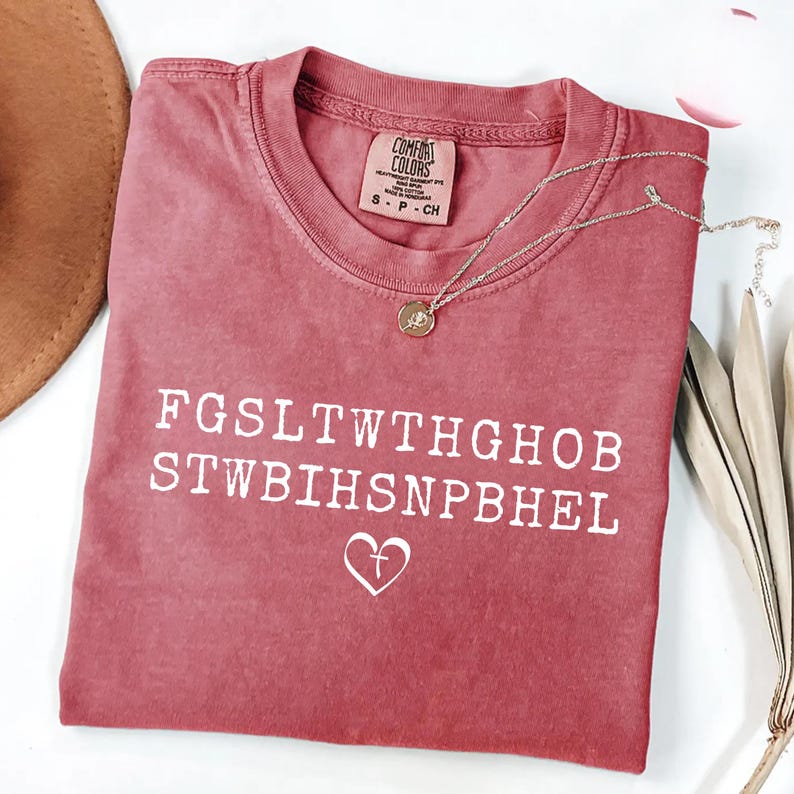 May include: A dusty rose Comfort Colors t-shirt with white text and a heart with a cross symbol. A gold necklace with a pendant rests on the shirt. The shirt is folded and has a tag.