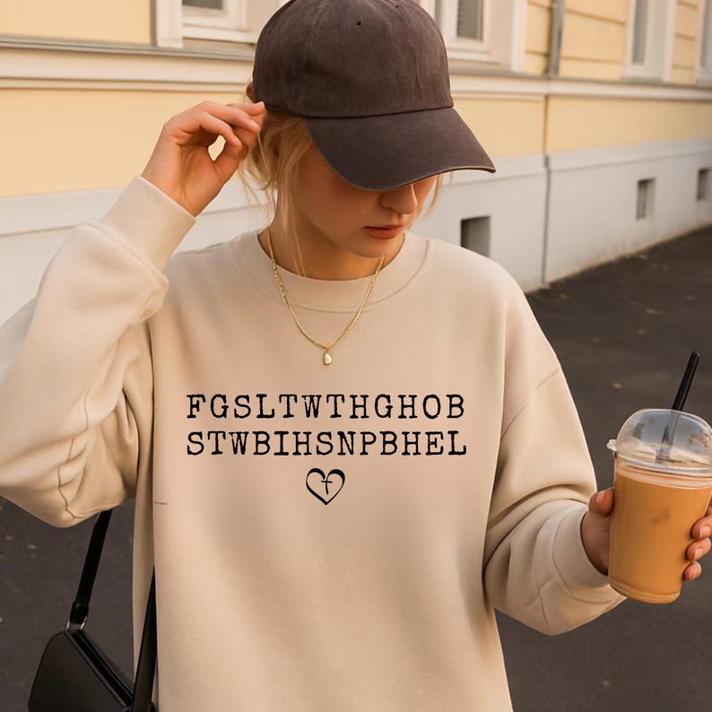 May include: A beige sweatshirt with the text "FGSLT WTHGHOB STWBIHSNPBHEL" and a heart symbol. The person is wearing a brown baseball cap and a gold necklace. A clear plastic cup with a brown beverage is held in the hand.