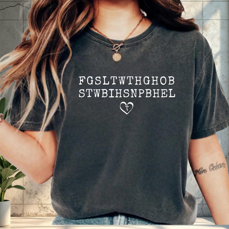 May include: Dark grey t-shirt featuring white text and a heart symbol. The text is a series of letters, with a heart and cross symbol below. The shirt has a crew neck and short sleeves.