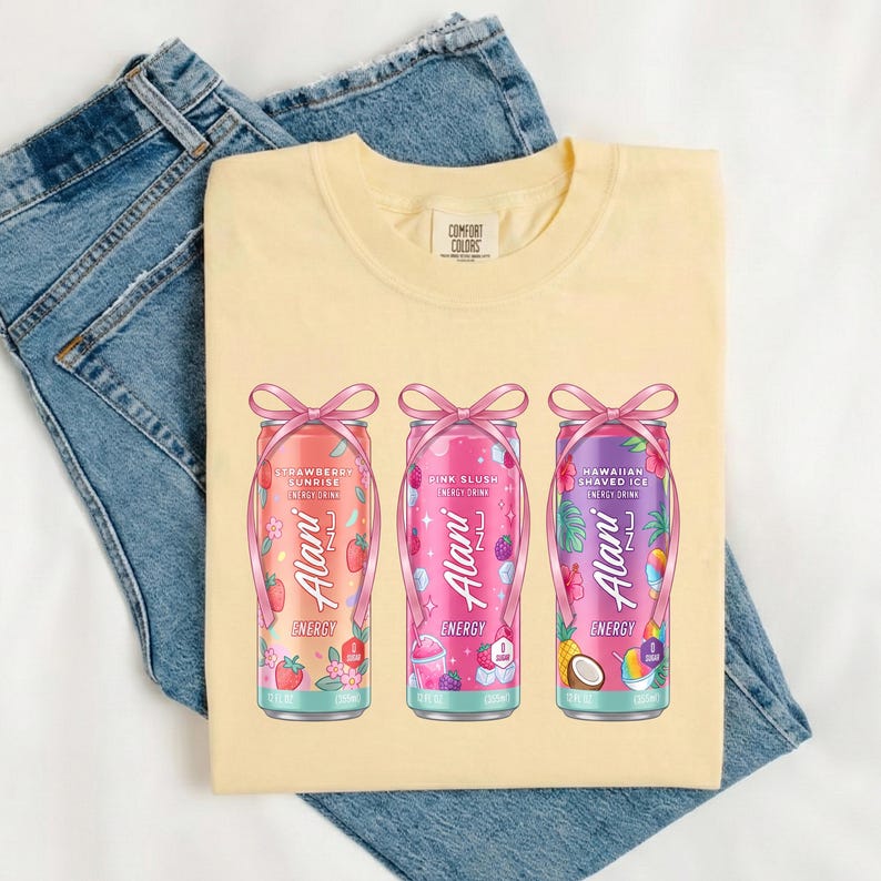 May include: A light yellow Comfort Colors t-shirt featuring three colorful Alani Nu energy drink cans with pink bows. The cans are Strawberry Sunrise, Pink Slush, and Hawaiian Shaved Ice flavors. The shirt is laid on top of a pair of blue jeans.