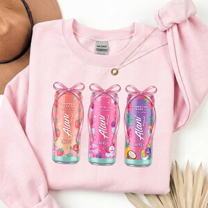 May include: A light pink sweatshirt featuring three Alani Nu energy drink cans with pink bows. The cans are decorated with fruit and floral designs, and the flavors are Strawberry Sunrise, Pink Slush, and Hawaiian Shaved Ice.