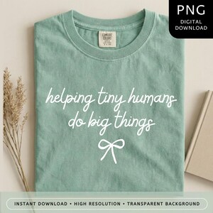 Può includere: Una t-shirt verde salvia con scollo rotondo ed etichetta Comfort Colors. La parte anteriore presenta una scritta bianca in corsivo che recita helping tiny humans do big things, posizionata sopra un semplice disegno a linee di un fiocco.