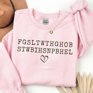 May include: A pale pink sweatshirt featuring the text "FGSLTWTHGHOB STWBIHSNPBHEL" in black, and a heart symbol. A gold necklace with a small pendant is draped over the sweatshirt.