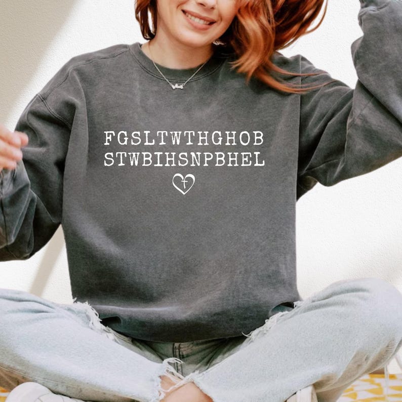 May include: A dark grey sweatshirt featuring white text and a heart-shaped cross graphic. The text is arranged in two lines, with the heart-shaped cross below. The sweatshirt has a vintage, washed appearance.