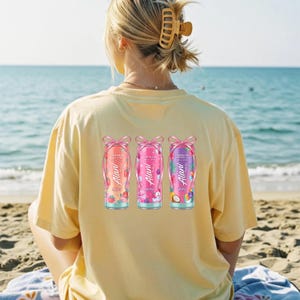 May include: A light yellow t-shirt with three colorful beverage can graphics on the back. Each can features a different fruit design and the text "Alani Nu Energy". The cans are adorned with pink bows.