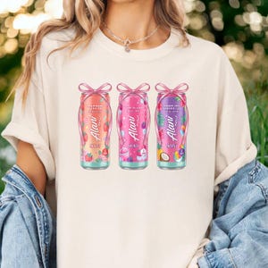 May include: A cream-colored t-shirt featuring three colorful energy drink cans. Each can is adorned with a pink bow and features the brand name "Alani Nu" and the flavors "Strawberry Sunrise", "Pink Slush", and "Hawaiian Shaved Ice".