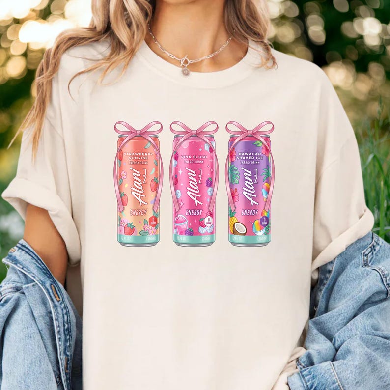 May include: A cream-colored t-shirt featuring three colorful energy drink cans. Each can is adorned with a pink bow and features the brand name "Alani Nu" and the flavors "Strawberry Sunrise", "Pink Slush", and "Hawaiian Shaved Ice".