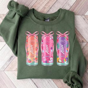 May include: A green sweatshirt featuring three colorful Alani Nu energy drink cans. The cans are decorated with pink bows and feature flavors like Strawberry Sunrise, Pink Slush, and Hawaiian Shaved Ice. The sweatshirt is a solid green color.