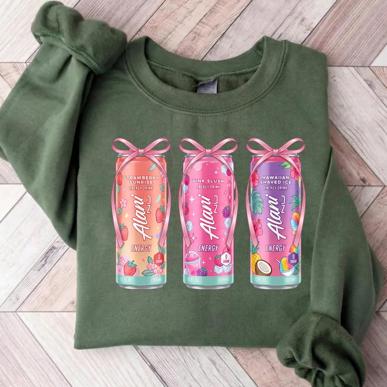 May include: A green sweatshirt featuring three colorful Alani Nu energy drink cans. The cans are decorated with pink bows and feature flavors like Strawberry Sunrise, Pink Slush, and Hawaiian Shaved Ice. The sweatshirt is a solid green color.