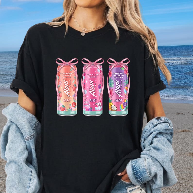 May include: Black t-shirt featuring three colorful energy drink cans with pink bows. The cans have the brand name "Alani" and various fruit illustrations. The flavors are Strawberry Sunrise, Pink Blush, and Hawaiian Shaved Ice.