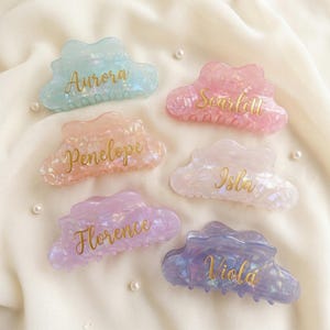 May include: Six cloud-shaped hair clips in pastel colors: light blue, pink, peach, white, lavender, and purple. Each clip has a name in gold script: Aurora, Scarlett, Penelope, Isla, Florence, and Viola. The clips are on a white fabric.