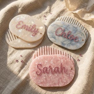 May include: Three personalized hair combs. One is off-white with the name "Emily" in pink rhinestones. Another is blue-grey with "Chloe". The third is pink with "Sarah". All have a matching comb base and are on a neutral fabric.