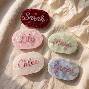 May include: Five oval-shaped name tags in shades of red, pink, green, white, and blue. Each tag features a different name spelled out in a cursive font with sparkling embellishments: Sarah, Lily, Maya, Chloe, and Grace.