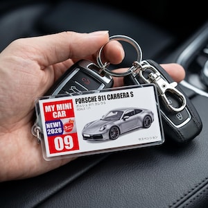 May include: A rectangular clear acrylic keyring featuring a graphic of a silver Porsche 911 Carrera S. The insert displays the text MY MINI CAR, NEW 2026, 09, PORSCHE 911 CARRERA S, and Japanese characters. It is attached to a metal split ring.