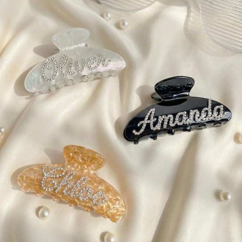 May include: Three claw hair clips in white pearlescent, solid black, and amber tortoiseshell patterns. Each clip features a name written in cursive script using small, clear rhinestones: Oliver on the white, Amanda on the black, and Chloe on the amber.