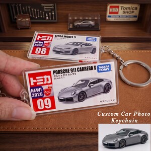 May include: A rectangular clear acrylic keychain featuring a printed insert of a silver Porsche 911 Carrera S, with the Takara Tomy logo, the number 09, and the text NEW! 2026. A metal split ring is attached to the side of the transparent plastic casing.
