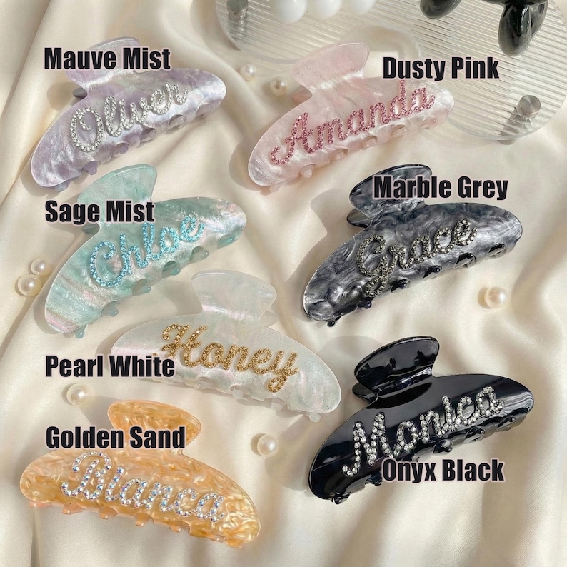 May include: A collection of seven acrylic hair claw clips in various colors including mauve mist, dusty pink, sage mist, marble grey, pearl white, golden sand, and onyx black. Each clip features a custom name spelled out in sparkling rhinestones.