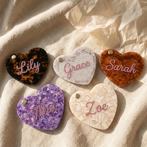 May include: Five heart-shaped personalized keychains. Each keychain features a different color and pattern, including tortoise shell, white, brown, purple, and cream. Each heart has a name spelled out in pink rhinestones: Lily, Grace, Sarah, Mia, and Zoe.