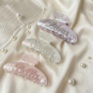May include: Three translucent hair claw clips in shades of lavender, iridescent white, and soft pink. Each clip features a name spelled out in small, clear rhinestones: Luna on the purple clip, Stella on the white clip, and Bella on the pink clip.