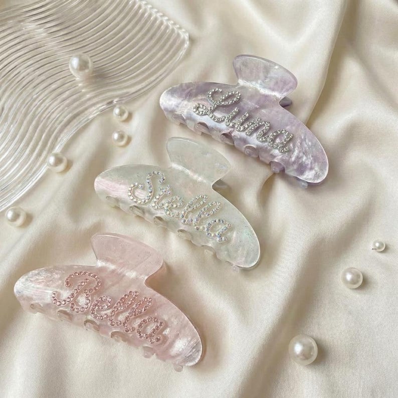 May include: Three translucent hair claw clips in shades of lavender, iridescent white, and soft pink. Each clip features a name spelled out in small, clear rhinestones: Luna on the purple clip, Stella on the white clip, and Bella on the pink clip.