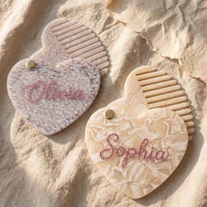 May include: Two heart-shaped hair combs with personalized names. One comb is light gray with the name "Olivia" in pink rhinestones. The other is cream-colored with the name "Sophia" in pink rhinestones. Both combs have a gold-colored stud.