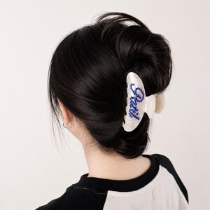 May include: A white, marbled-effect hair claw clip featuring the word "Paris" written in small, vibrant blue rhinestones. The clip is secured in dark hair, showcasing a curved, functional design with visible teeth for holding hair in place.