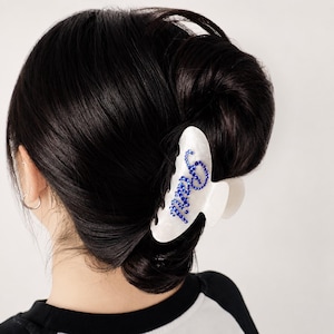 May include: A white pearlescent hair claw clip featuring the word "Bride" written in small, vibrant blue rhinestones. The clip is designed with a curved, multi-pronged structure to secure hair in place, showcasing a glossy finish and sparkling blue accents.