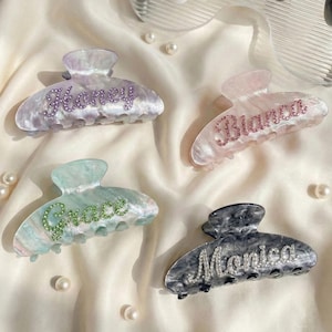 May include: Four marbled acrylic hair claw clips featuring personalized names in sparkling rhinestones. The clips include the names Honey in purple, Bianca in pink, Grace in green, and Monica in clear crystals, each set against a unique color.