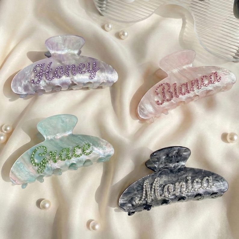 May include: Four marbled acrylic hair claw clips featuring personalized names in sparkling rhinestones. The clips include the names Honey in purple, Bianca in pink, Grace in green, and Monica in clear crystals, each set against a unique color.