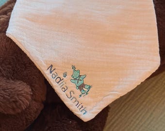 May include: A brown teddy bear with a white bib featuring the name "Nadia Smith" and a small illustration. The bib is resting on the bear's chest. The bear is lying on a light blue surface.