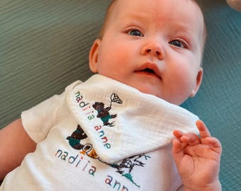 May include: A baby wearing a white onesie and bib with the embroidered text "nadiia anne" and a graphic of a bear catching fish. A plush lamb toy rests above the baby on a teal surface.