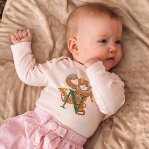 May include: A baby wearing a light pink long-sleeved bodysuit with a cross-stitched design of a bear holding a toy and the letter "N". The baby is also wearing pink corduroy shorts with a bow.