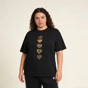 May include: Black t-shirt featuring a vertical arrangement of gold-colored heart designs. The hearts vary in pattern, including a crown, leopard print, and solid outlines. The shirt has short sleeves and a crew neck.