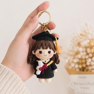 May include: Crocheted graduation keychain featuring a doll in a black cap and gown with a gold tassel. The doll holds a diploma tied with a red ribbon. The keychain has a gold ring.