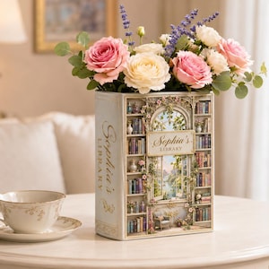 Peut inclure: Un vase décoratif en forme de livre, rempli de roses roses et crème, de lavande et de verdure. Le vase est orné d'une scène de bibliothèque et du texte "Sophia's Library". Une tasse et une soucoupe sont posées sur une table ronde.