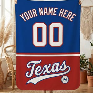 Może przedstawiać: Niebiesko-czerwony koc z napisem "YOUR NAME HERE" i numerem "00" w kolorze białym. Dolna część zawiera napis "Texas" w czcionce skryptowej z grafiką baseballową. Koc jest trzymany przez osobę.