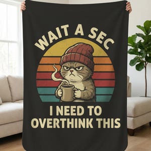 Op de afbeelding: Zwarte deken met een chagrijnige cartoonkat met een rode muts en een koffiemok. De deken heeft de tekst "WAIT A SEC I NEED TO OVERTHINK THIS" in witte letters tegen een retro zonsondergang achtergrond.