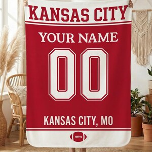 Pode incluir: Cobertor vermelho e branco com o texto "KANSAS CITY" no topo, "YOUR NAME" no meio e "00" em grandes números brancos. A parte inferior diz "KANSAS CITY, MO" com um gráfico de futebol. Um cobertor personalizado com tema esportivo.