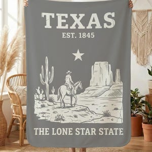 May include: Grey blanket with a white graphic of a cowboy on horseback in a desert scene, with the text "TEXAS EST. 1845" and "THE LONE STAR STATE". The blanket is being held up by a person.