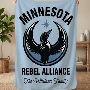 May include: A light blue blanket with the word "MINNESOTA" above a black bird emblem with outstretched wings, a star, and the words "REBEL ALLIANCE" and "The Williams Family" in black text. The blanket is held up by two hands.