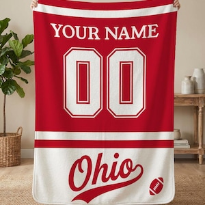 May include: Red and white personalized football jersey blanket. The blanket features the text "YOUR NAME" above the number "00" and the word "Ohio" with a football graphic. The blanket is held up by two hands.