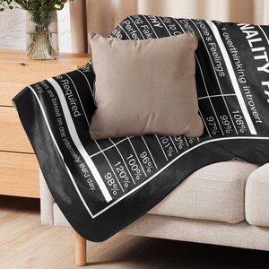May include: A black throw blanket with white text and a beige pillow on a sofa. The blanket has a chart-like design with percentages and phrases like "overthinking introvert." The pillow is square and the sofa has wooden legs.