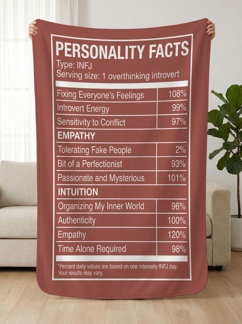 May include: A maroon blanket with white text that reads "PERSONALITY FACTS." The text lists personality traits with percentages, including "Fixing Everyone's Feelings," "Introvert Energy," and "Time Alone Required." The blanket is held up by hands.